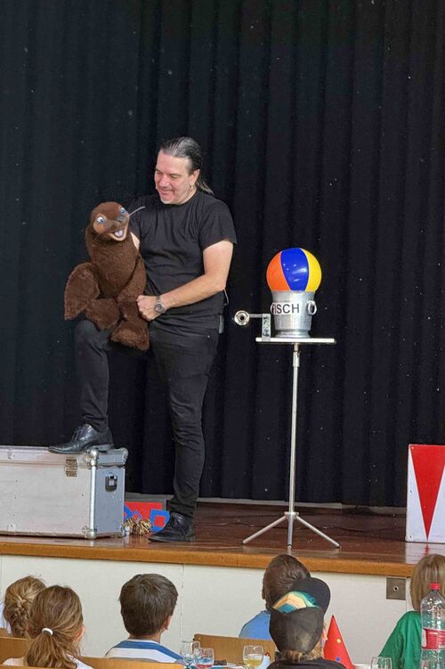 Ein Zauberer steht auf der Bühne und hat ein Stofftier in der Hand. Er redet mit dem Bauch.