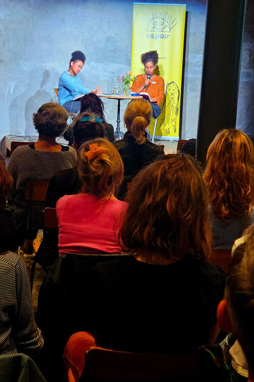 Im Vordergrund sitzt das Publikum der Lesung, im Hintergrund sieht man Fatima Moumouni und Anja Glover, die aus ihrem Buch liest.