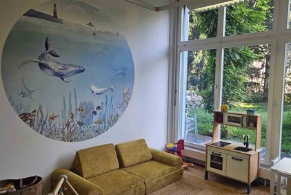 Das Bild zeigt ein Besuchszimmer mit einem gemütlichen, gelben Sofa, einem Wandbild mit Meerestieren sowie einer Spielküche.